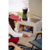 Personne portant une casquette rose et un haut blanc atteint un compartiment gris en plastique ouvert avec des emballages de snacks visibles (sachet bleu) ; une cafetière noire est posée sur la boîte, une chaise pliante bleue à côté, de l'herbe en arrière-plan