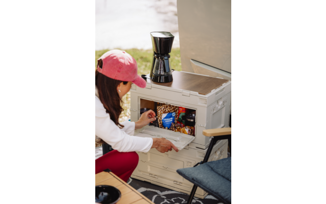 Personne portant une casquette rose et un haut blanc atteint un compartiment gris en plastique ouvert avec des emballages de snacks visibles (sachet bleu) ; une cafetière noire est posée sur la boîte, une chaise pliante bleue à côté, de l'herbe en arrière-plan