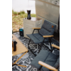 Deux chaises pliantes rembourrées avec accoudoirs en bois sur un tapis d'extérieur à motif à côté d'une portière de véhicule ouverte ; une caisse plastique empilable avec une cafetière moka, un petit appareil d'alimentation bleu-noir, une table en bois avec un mug de voyage ; rive herbeuse en arrière-plan