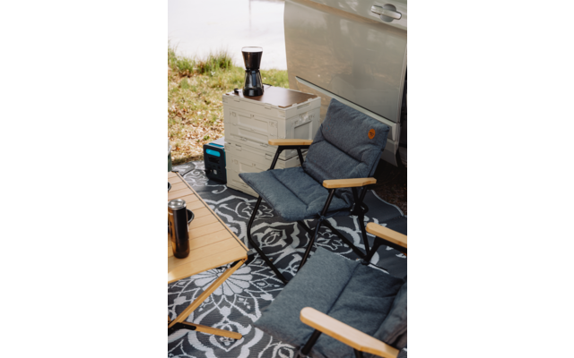 Deux chaises pliantes rembourrées avec accoudoirs en bois sur un tapis d'extérieur à motif à côté d'une portière de véhicule ouverte ; une caisse plastique empilable avec une cafetière moka, un petit appareil d'alimentation bleu-noir, une table en bois avec un mug de voyage ; rive herbeuse en arrière-plan