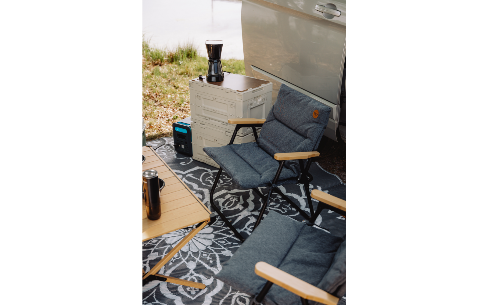 Deux chaises pliantes rembourrées avec accoudoirs en bois sur un tapis d'extérieur à motif à côté d'une portière de véhicule ouverte ; une caisse plastique empilable avec une cafetière moka, un petit appareil d'alimentation bleu-noir, une table en bois avec un mug de voyage ; rive herbeuse en arrière-plan