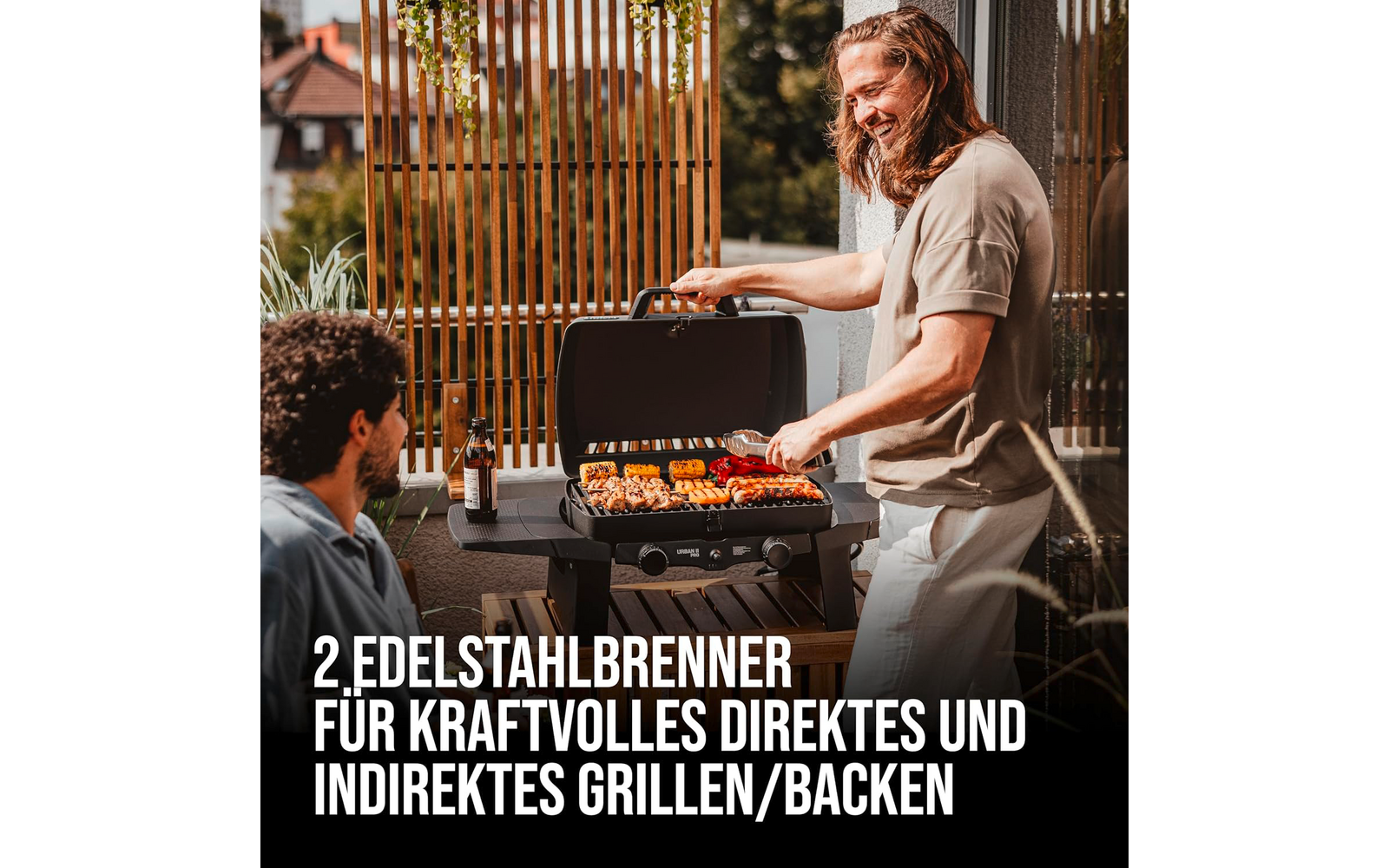 Two men on a balcony; one stands at a grill turning skewers of vegetables and meat, the other sits nearby with a beer bottle