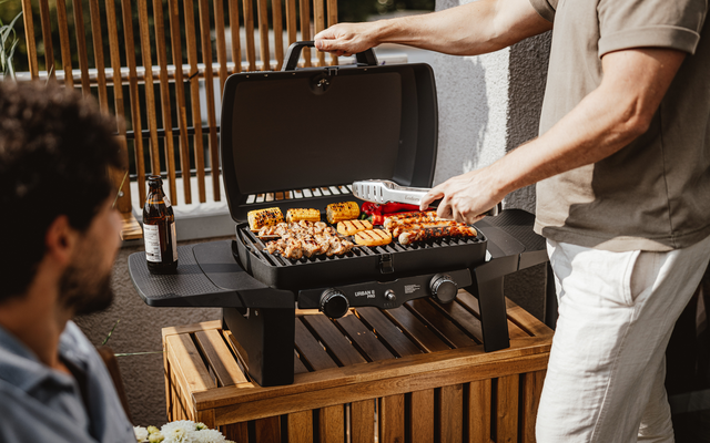 Two people at a portable grill; standing person flips skewers, sausages, and corn; beer bottle on table; wooden slatted background