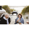 Two people sit on the beach at a table with food and drinks; on the left a man wearing a baseball cap, on the right a woman with a yellow headband; behind them a portable grill on a stand, sand dunes and the beach in the background; rear of a vehicle visible at left