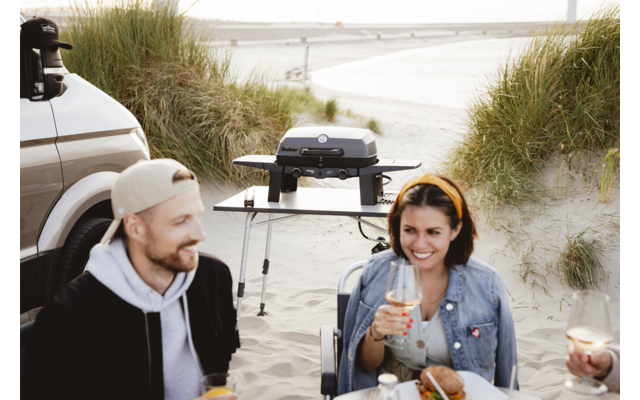 Two people sit on the beach at a table with food and drinks; on the left a man wearing a baseball cap, on the right a woman with a yellow headband; behind them a portable grill on a stand, sand dunes and the beach in the background; rear of a vehicle visible at left