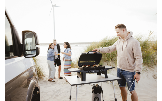 Person operating a portable grill on a folding table at a sandy beach; three people stand in the background next to a white camper van, sand dunes and a wind turbine visible