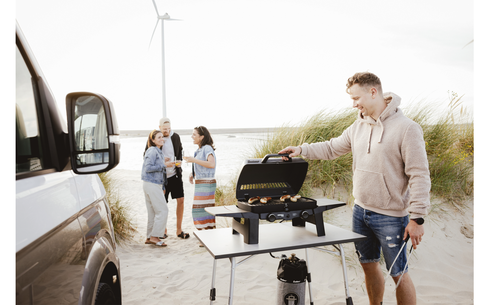 Person operating a portable grill on a folding table at a sandy beach; three people stand in the background next to a white camper van, sand dunes and a wind turbine visible