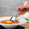 Hand mit rot lackierten Fingernägeln hält einen Metalllöffel über einer weißen Schale mit orangefarbener Suppe und drei Croutons auf einem Holztisch vor grauem Hintergrund