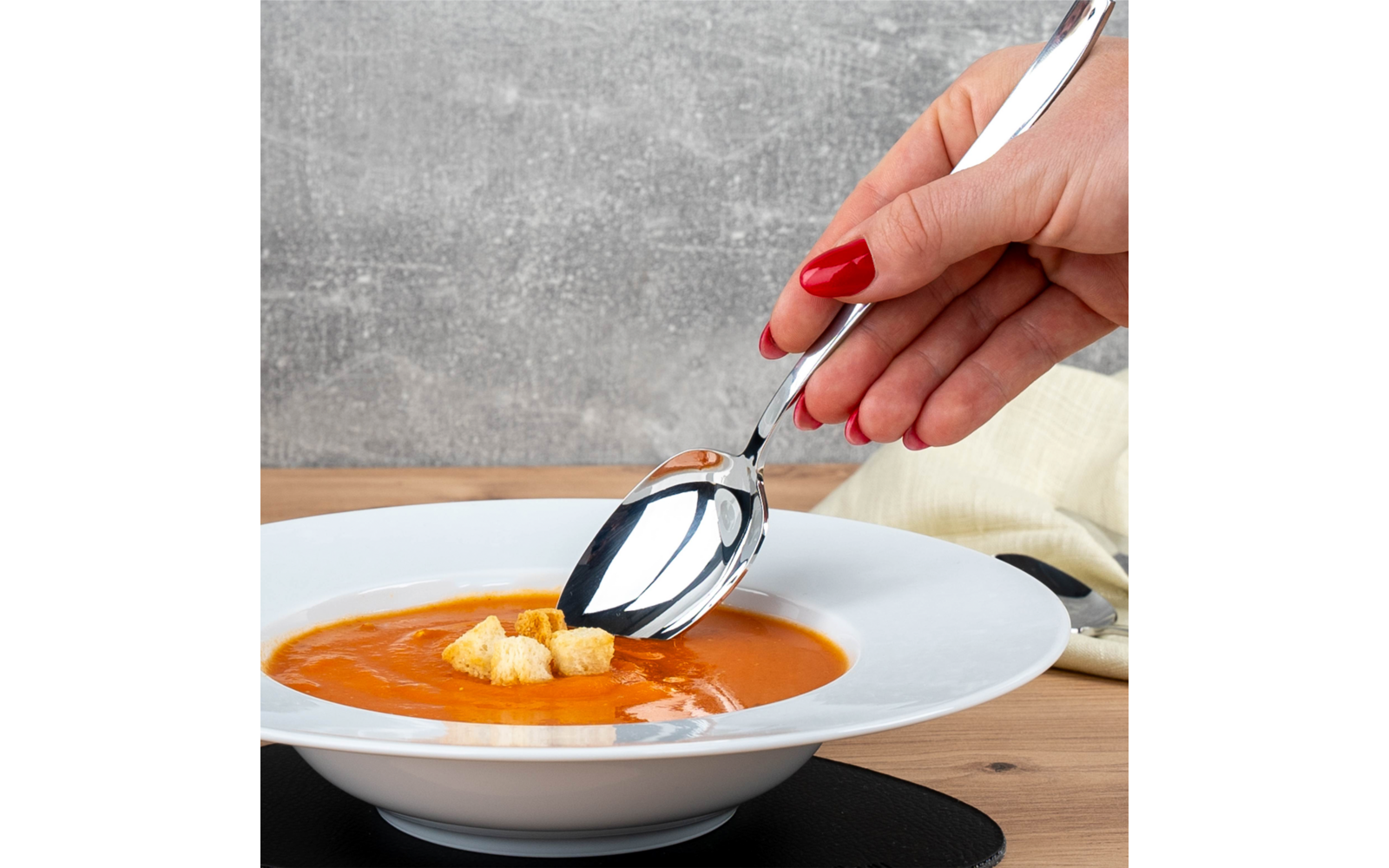 Hand mit rot lackierten Fingernägeln hält einen Metalllöffel über einer weißen Schale mit orangefarbener Suppe und drei Croutons auf einem Holztisch vor grauem Hintergrund