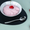 Weiße Schale mit rosa cremiger Masse und einer roten Kirsche obenauf, auf rundem schwarzem Untersetzer, silberner Löffel davor, am rechten Bildrand einzelne Kirschen sichtbar