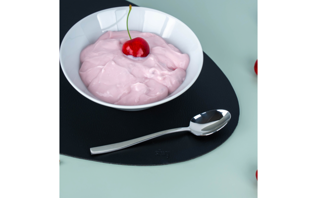Weiße Schale mit rosa cremiger Masse und einer roten Kirsche obenauf, auf rundem schwarzem Untersetzer, silberner Löffel davor, am rechten Bildrand einzelne Kirschen sichtbar