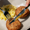 Beiger Teller mit zwei runden, braunen Lebensmittelstücken, Kartoffelsalat mit heller Sauce und einem kleinen Petersilienblatt; Hand hält Messer und Gabel und schneidet eines der Stücke