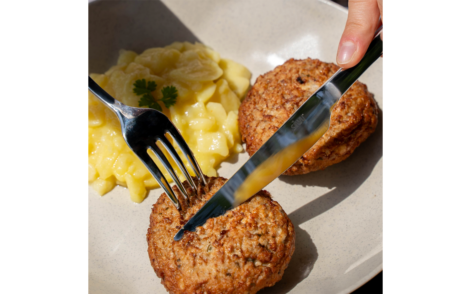 Beiger Teller mit zwei runden, braunen Lebensmittelstücken, Kartoffelsalat mit heller Sauce und einem kleinen Petersilienblatt; Hand hält Messer und Gabel und schneidet eines der Stücke