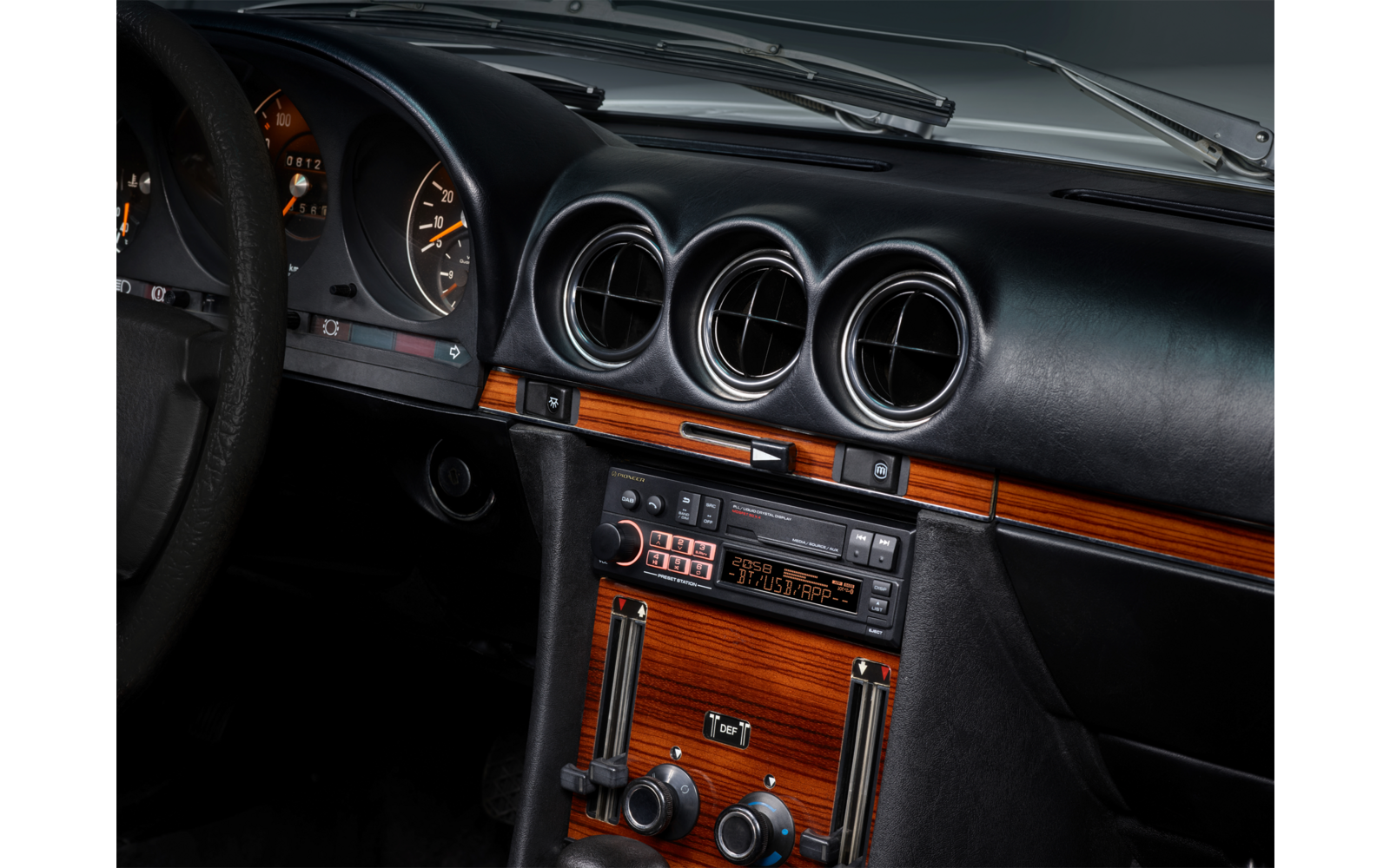 Innenraum eines Autos: schwarzes Armaturenbrett mit drei runden Lüftungsdüsen, Holzblende, Radio mit Kassettenschacht und Tasten, Klimabedienelemente, Teil des Lenkrads und Instrumente, Windschutzscheibe mit Wischer