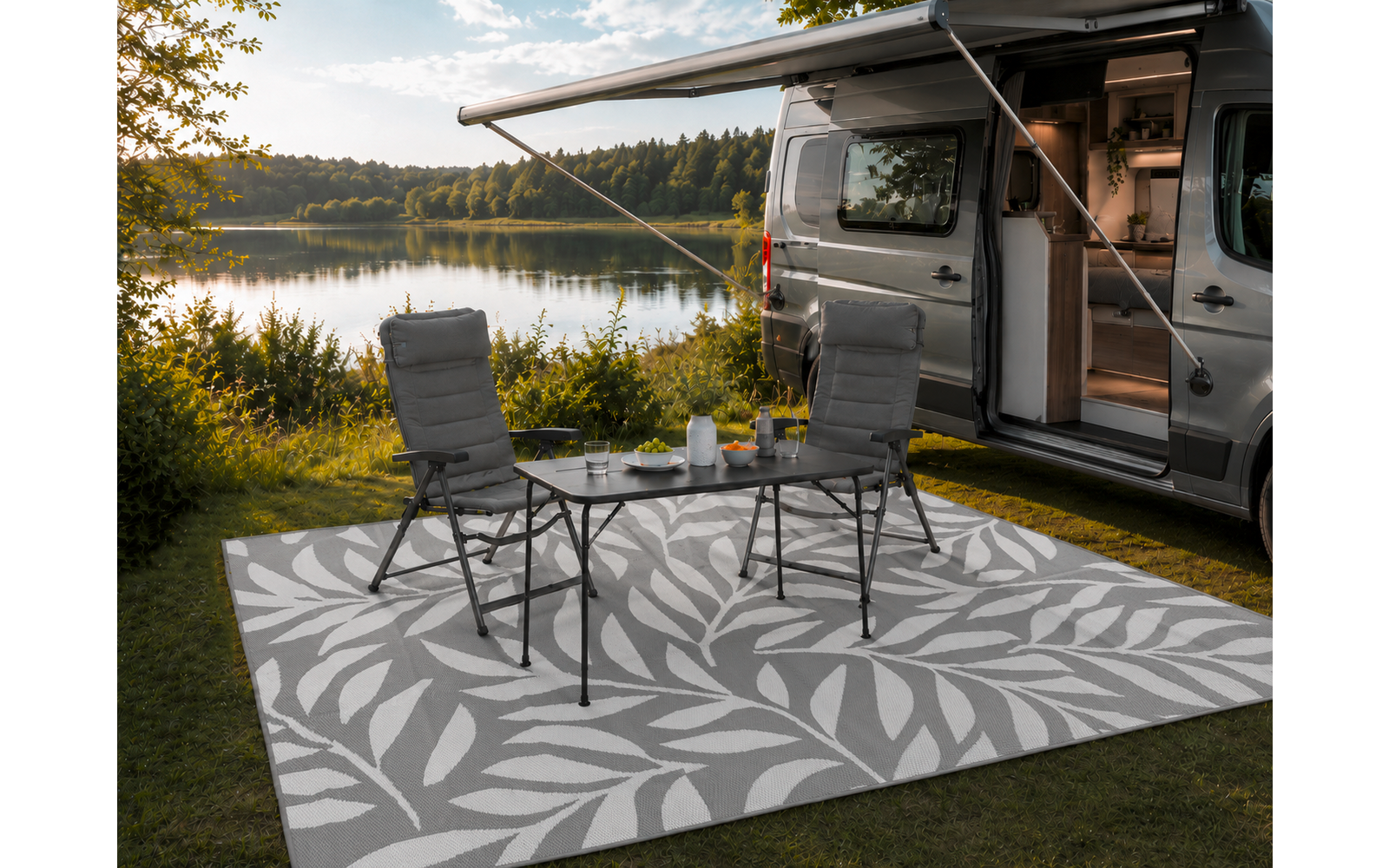 Wohnmobil am Ufer eines Sees, Schiebetür geöffnet. Zwei Campingstühle und kleiner Klapptisch auf einem hell gemusterten Außen­teppich. Auf dem Tisch Teekanne und Tassen. Gras, Bäume, See im Hintergrund, wolkiger Himmel
