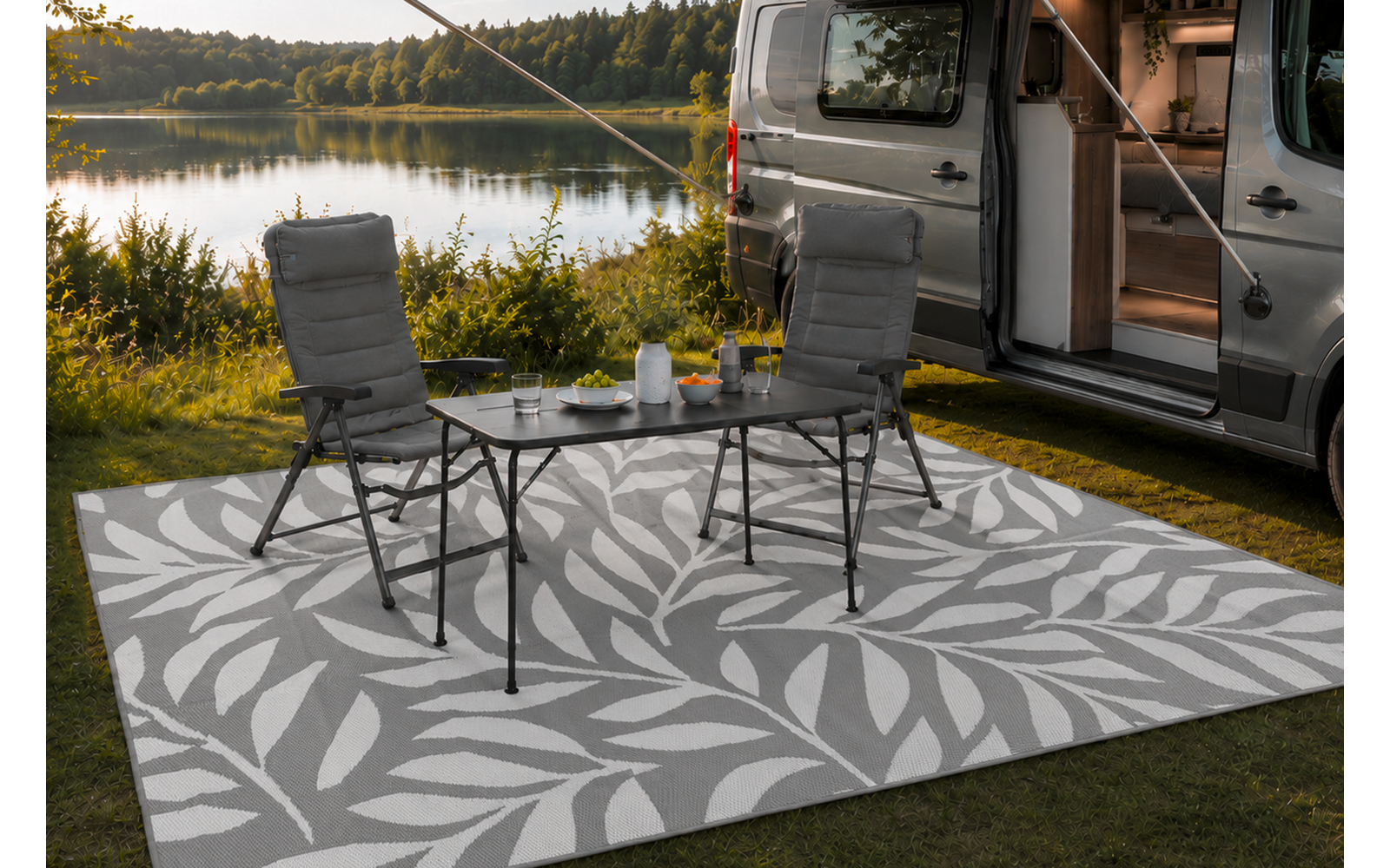 Camper geparkeerd aan de oever van een meer, schuifdeur open. Twee campingstoelen en een klein inklapbaar tafeltje op een licht patroon buitenkleed. Theepot en kopjes op de tafel. Gras, bomen en meer op de achtergrond, bewolkte lucht