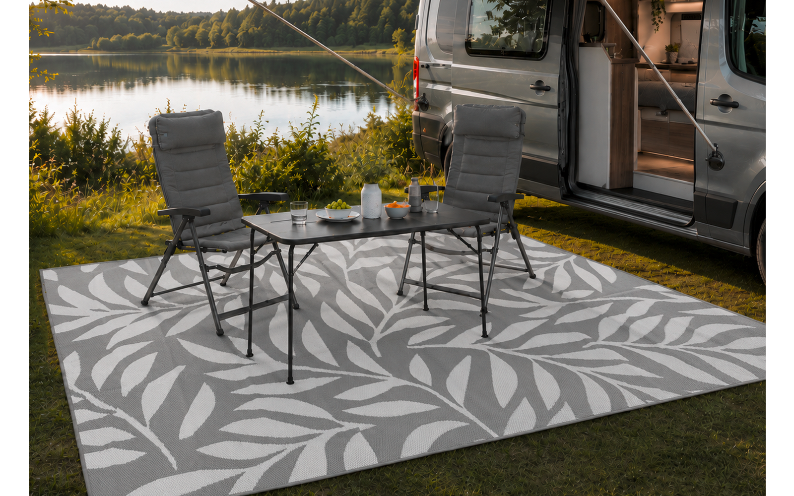 Wohnmobil am Ufer eines Sees, Schiebetür geöffnet. Zwei Campingstühle und kleiner Klapptisch auf einem hell gemusterten Außen­teppich. Auf dem Tisch Teekanne und Tassen. Gras, Bäume, See im Hintergrund, wolkiger Himmel