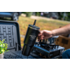 Hand holding a black tumbler with a straw and mountain logo in front of a camping stove on a folding table; small potted plant at left; blurred grassy background