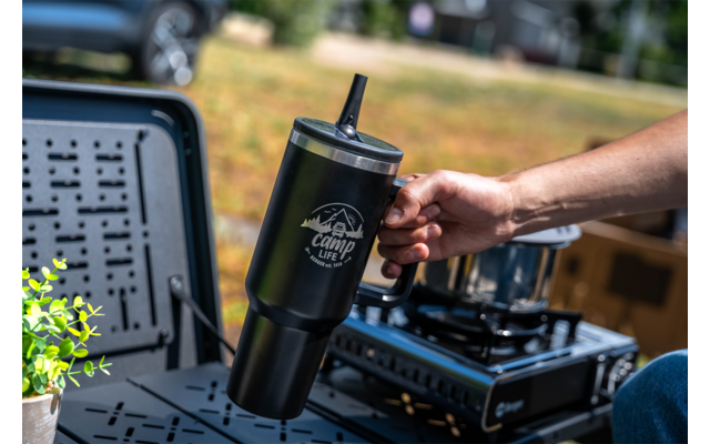 Hand holding a black tumbler with a straw and mountain logo in front of a camping stove on a folding table; small potted plant at left; blurred grassy background