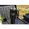 Black insulated travel mug with handle and silver lid, white 'camp life' logo with mountain graphic, on a black perforated metal table; small green plant left, blurred camping stove right, blurred grassy background