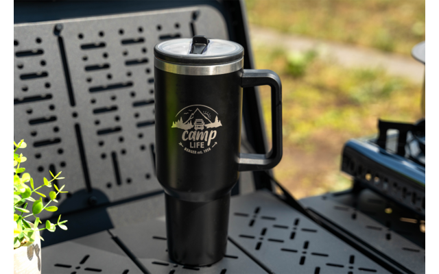Black insulated travel mug with handle and silver lid, white 'camp life' logo with mountain graphic, on a black perforated metal table; small green plant left, blurred camping stove right, blurred grassy background