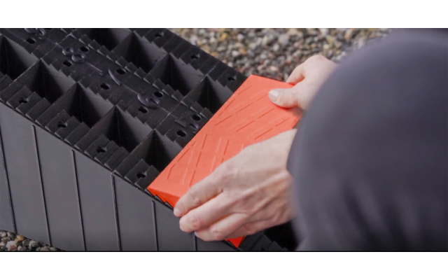 Deux mains tiennent un module carré orange au‑dessus d'une grille en plastique noire sur du gravier