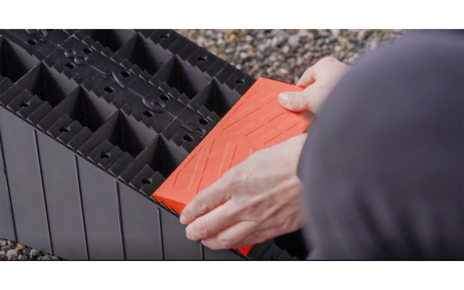 Deux mains tiennent un module carré orange au‑dessus d'une grille en plastique noire sur du gravier