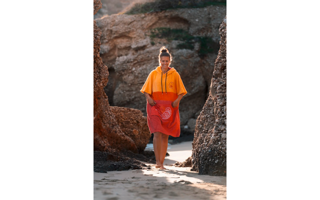 Person mit hochgesteckten Haaren, trägt ein orange-rotes Kapuzenhandtuch mit weißem kreisförmigem Muster, geht barfuß auf Sand zwischen Felsen