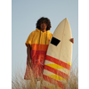 Person mit lockigem Haar trägt einen gelb-roten Kapuzenponcho und hält ein weißes Surfboard mit roten und gelben Streifen, steht in hohem Gras vor klarem Himmel