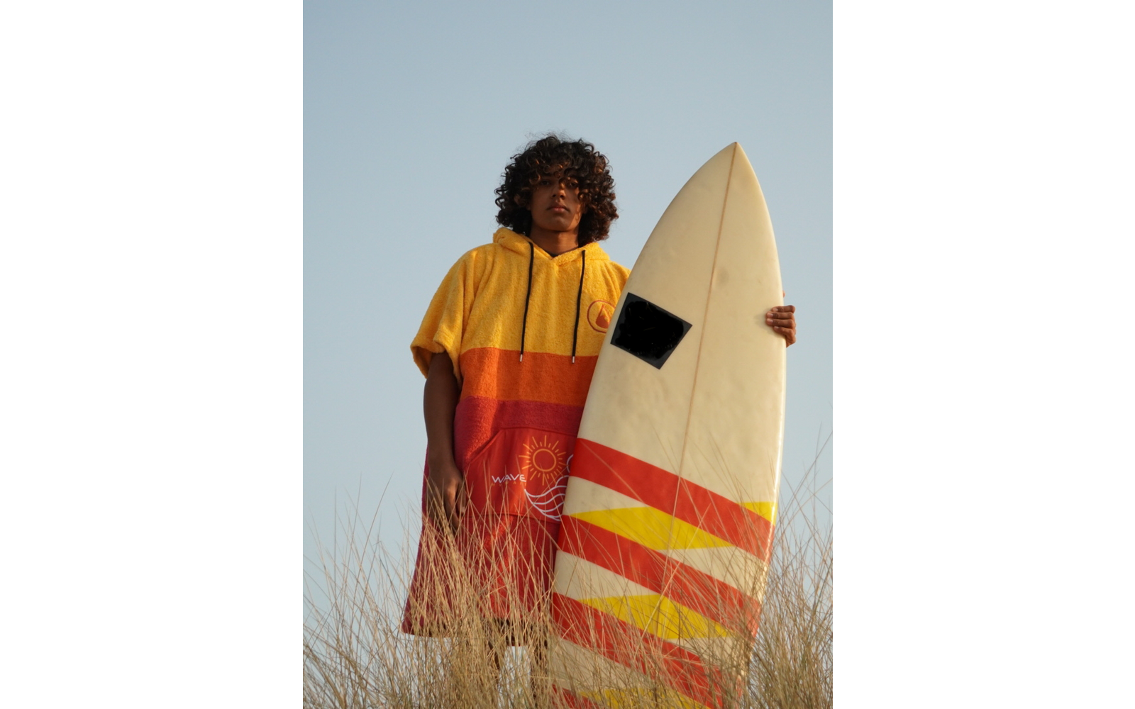 Person mit lockigem Haar trägt einen gelb-roten Kapuzenponcho und hält ein weißes Surfboard mit roten und gelben Streifen, steht in hohem Gras vor klarem Himmel