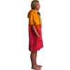 Person in Seitenansicht, steht barfuß mit Händen in den Taschen, trägt ein Kapuzenhandtuch/Poncho mit gelben, orange- und roten horizontalen Farbfeldern und weißer Aufdruck vorne unten
