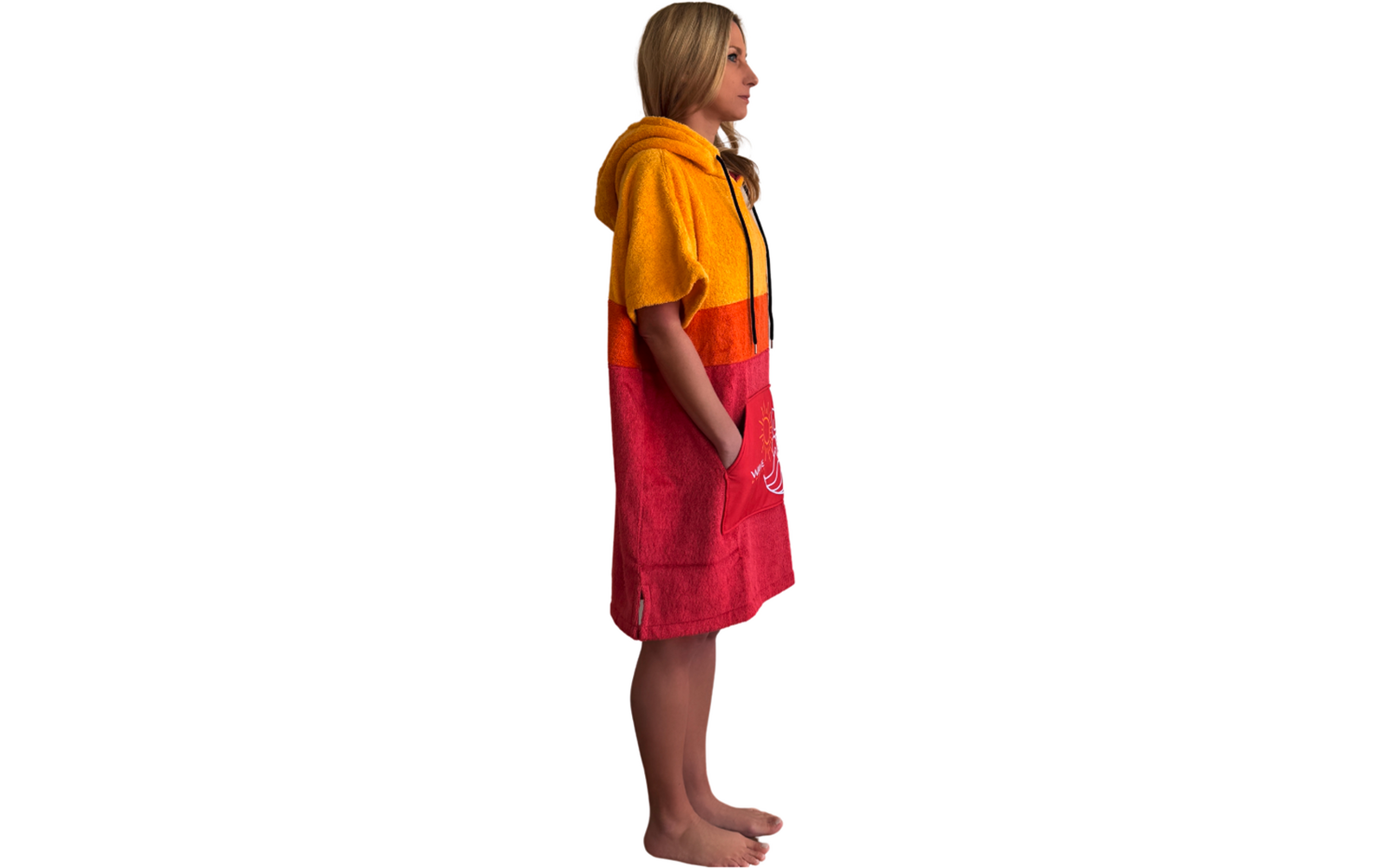 Person in Seitenansicht, steht barfuß mit Händen in den Taschen, trägt ein Kapuzenhandtuch/Poncho mit gelben, orange- und roten horizontalen Farbfeldern und weißer Aufdruck vorne unten