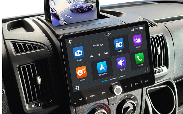 Console centrale d'un véhicule avec un grand écran tactile affichant des icônes d'applications, un écran relevable au‑dessus montrant une image d'une voiture garée et son environnement, bouches d'aération et commandes rotatives de climatisation