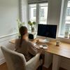 Person mit geflochtenem Haar sitzt in einem Sessel an einem Holztisch vor einem Computerbildschirm, Fenster im Hintergrund, mehrere Topfpflanzen, kleines grünes Gerät, Kaffeetasse und Glas mit Strohhalm auf dem Tisch