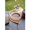 On a gray surface: a coiled orange gas hose with a brass connector and red shut-off valve attached to a thin braided metal line; in the background a green gas cylinder with a regulator, to the right a black gas canister with orange lettering, left a metal part of a stove; grass at the top of the image