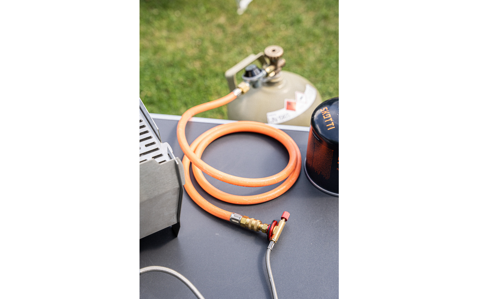 On a gray surface: a coiled orange gas hose with a brass connector and red shut-off valve attached to a thin braided metal line; in the background a green gas cylinder with a regulator, to the right a black gas canister with orange lettering, left a metal part of a stove; grass at the top of the image