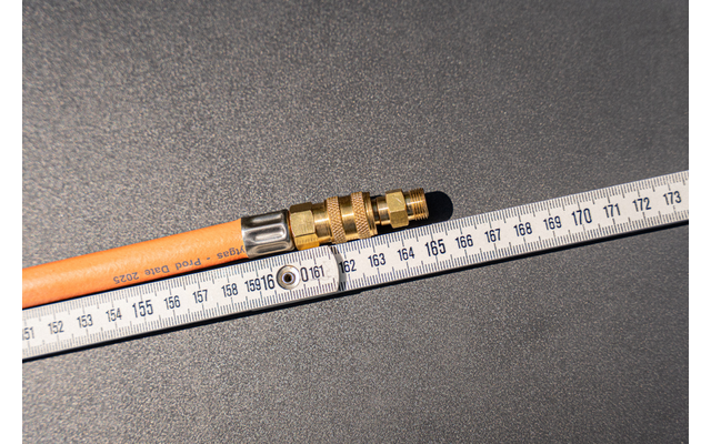Orange hose with brass fitting lies diagonally on a dark textured surface; a white folding ruler with centimeter markings lies beside it, numbers visible