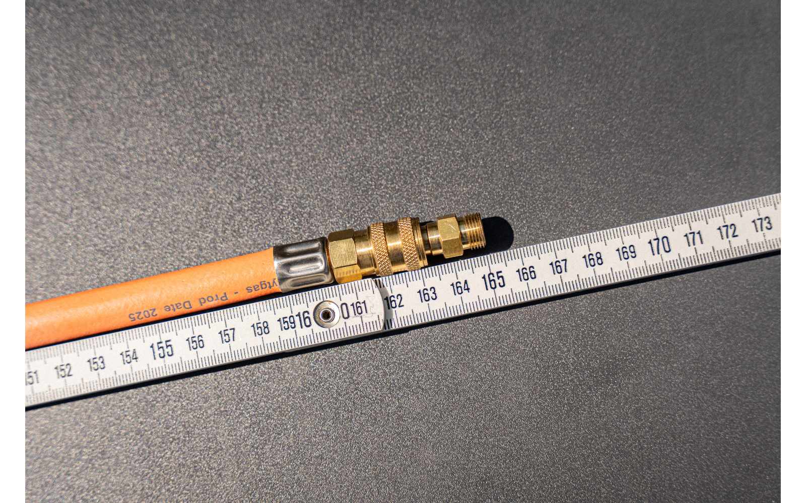 Orange hose with brass fitting lies diagonally on a dark textured surface; a white folding ruler with centimeter markings lies beside it, numbers visible