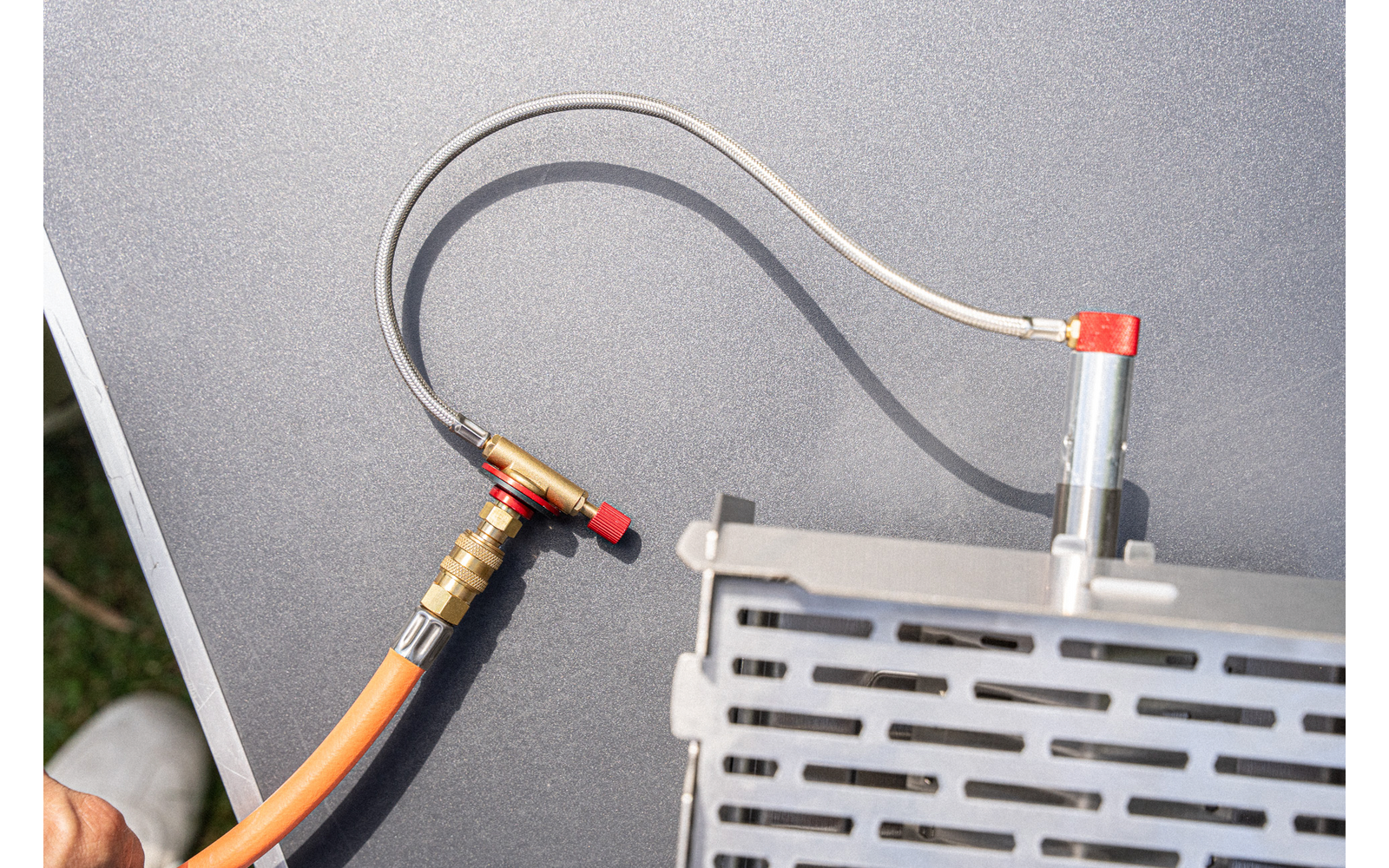 Orange hose with brass fitting and gray flexible tube connected to a metal device with perforated cover; dark matte surface in the background; hand holding the hose at bottom left