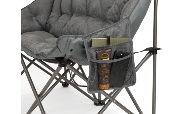 Grauer gepolsterter Klappstuhl mit Metallgestell und seitlicher Stofftasche mit mehreren Fächern; darin ein beiges flaches rechteckiges Objekt, ein schwarzer Stift, ein schwarzes Smartphone und ein zylindrischer schwarzer Behälter mit orangefarbenem 