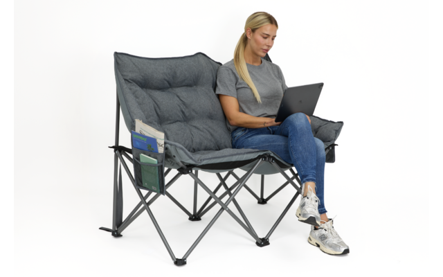 Frau sitzt auf grauem, klappbarem Doppel-Campingstuhl, Laptop auf dem Schoß, trägt graues T-Shirt, blaue Jeans und weiße Sneakers; Stuhl mit seitlichen Taschen und Zeitschriften; weißer Hintergrund