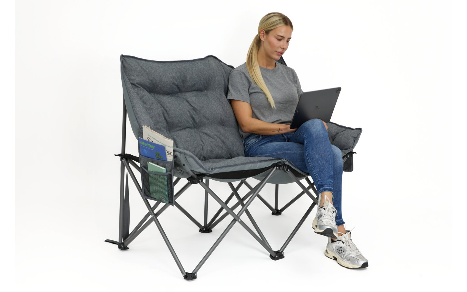 Frau sitzt auf grauem, klappbarem Doppel-Campingstuhl, Laptop auf dem Schoß, trägt graues T-Shirt, blaue Jeans und weiße Sneakers; Stuhl mit seitlichen Taschen und Zeitschriften; weißer Hintergrund