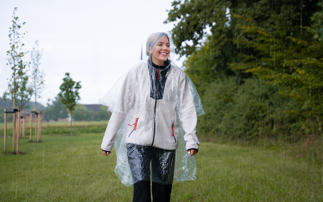 Personne aux cheveux gris courts porte une veste claire et un poncho de pluie transparent à capuche, marche sur une pelouse à côté d'arbres