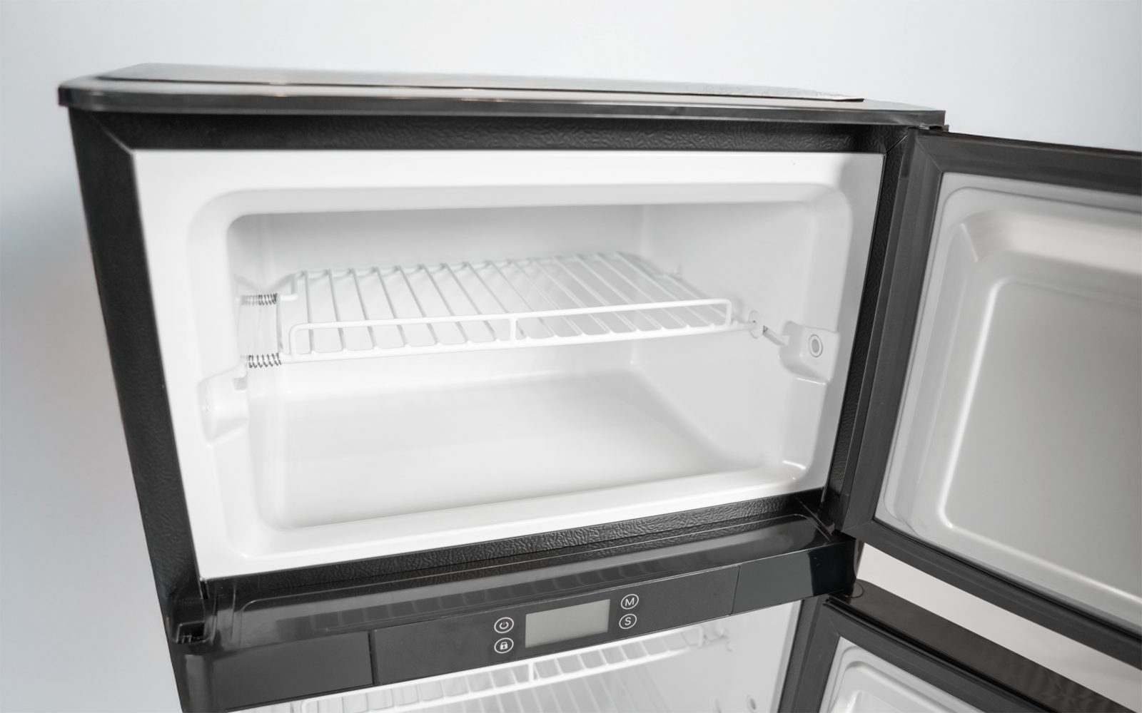 Offenes, leeres Gefrierfach eines Kühlschranks mit Drahtgitterablage und schwarzem Gehäuse