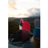 Person mit rosa Mütze und weißer Weste hält ein rotes Brett neben einem offenen Zelt; eine weitere Person teilweise im Vordergrund sichtbar; hügelige Landschaft und wolkiger Himmel mit Licht am Horizont