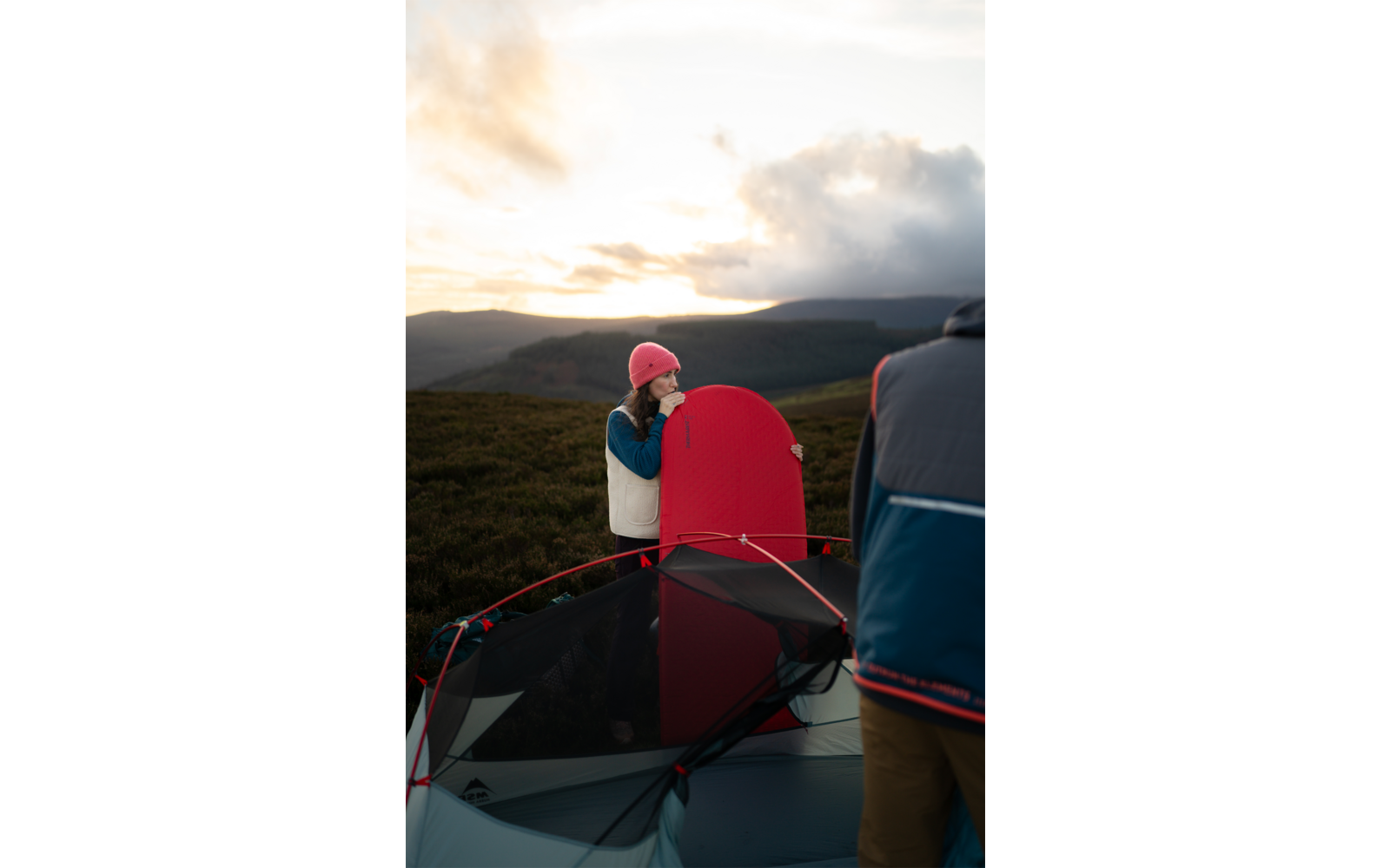 Person mit rosa Mütze und weißer Weste hält ein rotes Brett neben einem offenen Zelt; eine weitere Person teilweise im Vordergrund sichtbar; hügelige Landschaft und wolkiger Himmel mit Licht am Horizont