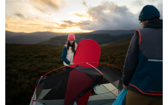Zwei Personen neben einem teilweise aufgebauten roten Zelt in hügeliger Landschaft bei Sonnenuntergang; eine Person mit rosa Mütze hält Zeltstoff, die andere trägt eine blaue Tasche