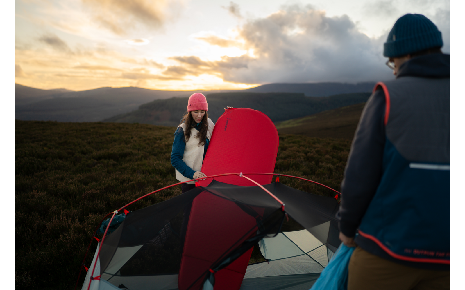 Zwei Personen neben einem teilweise aufgebauten roten Zelt in hügeliger Landschaft bei Sonnenuntergang; eine Person mit rosa Mütze hält Zeltstoff, die andere trägt eine blaue Tasche