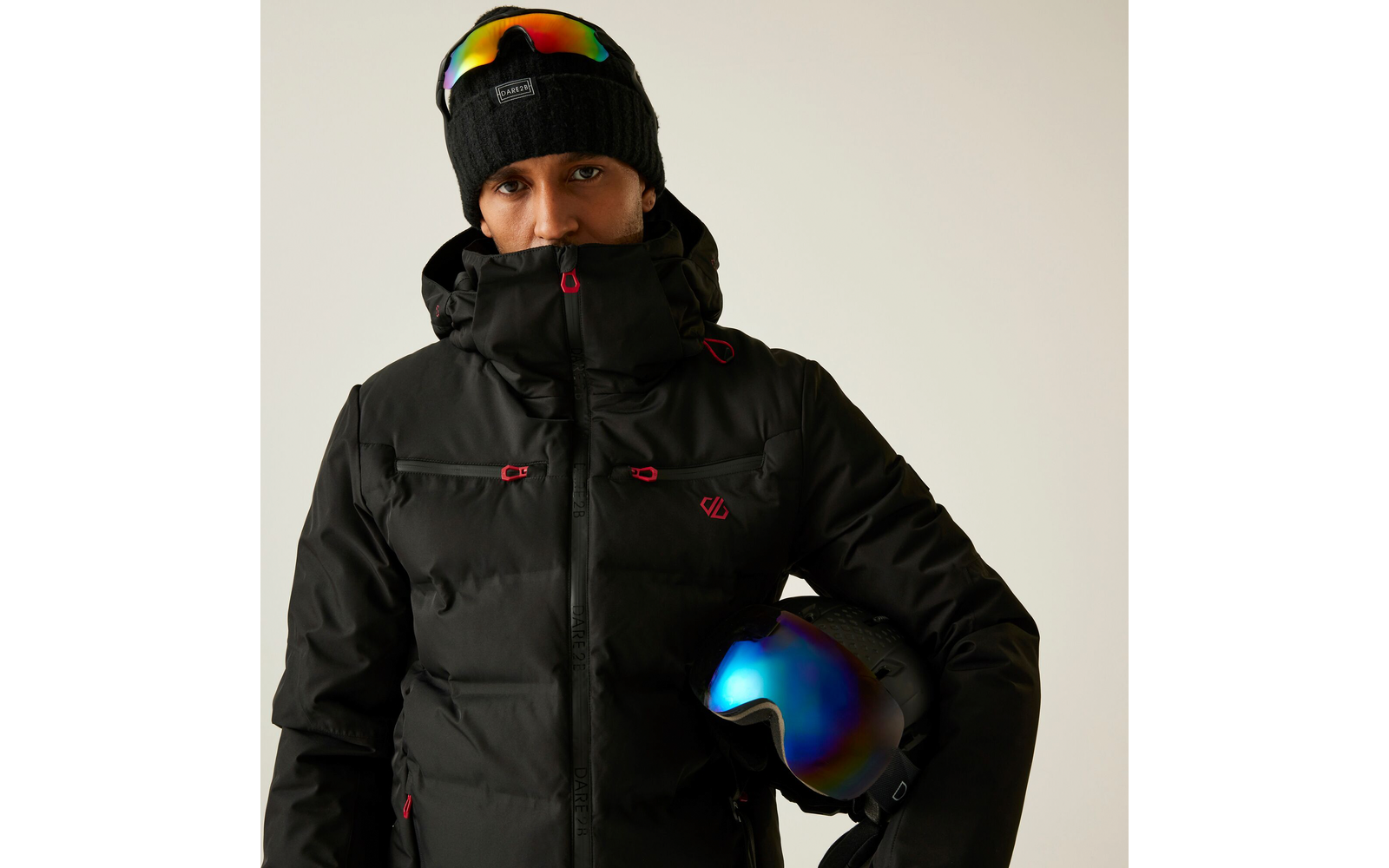 Person mit schwarzer Winterjacke, Kapuze und Skibrille auf dem Kopf, hält einen Skihelm unter dem Arm