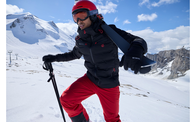 Person in roter Skihose und Helm mit Skistöcken auf schneebedecktem Berg bei Sonnenschein